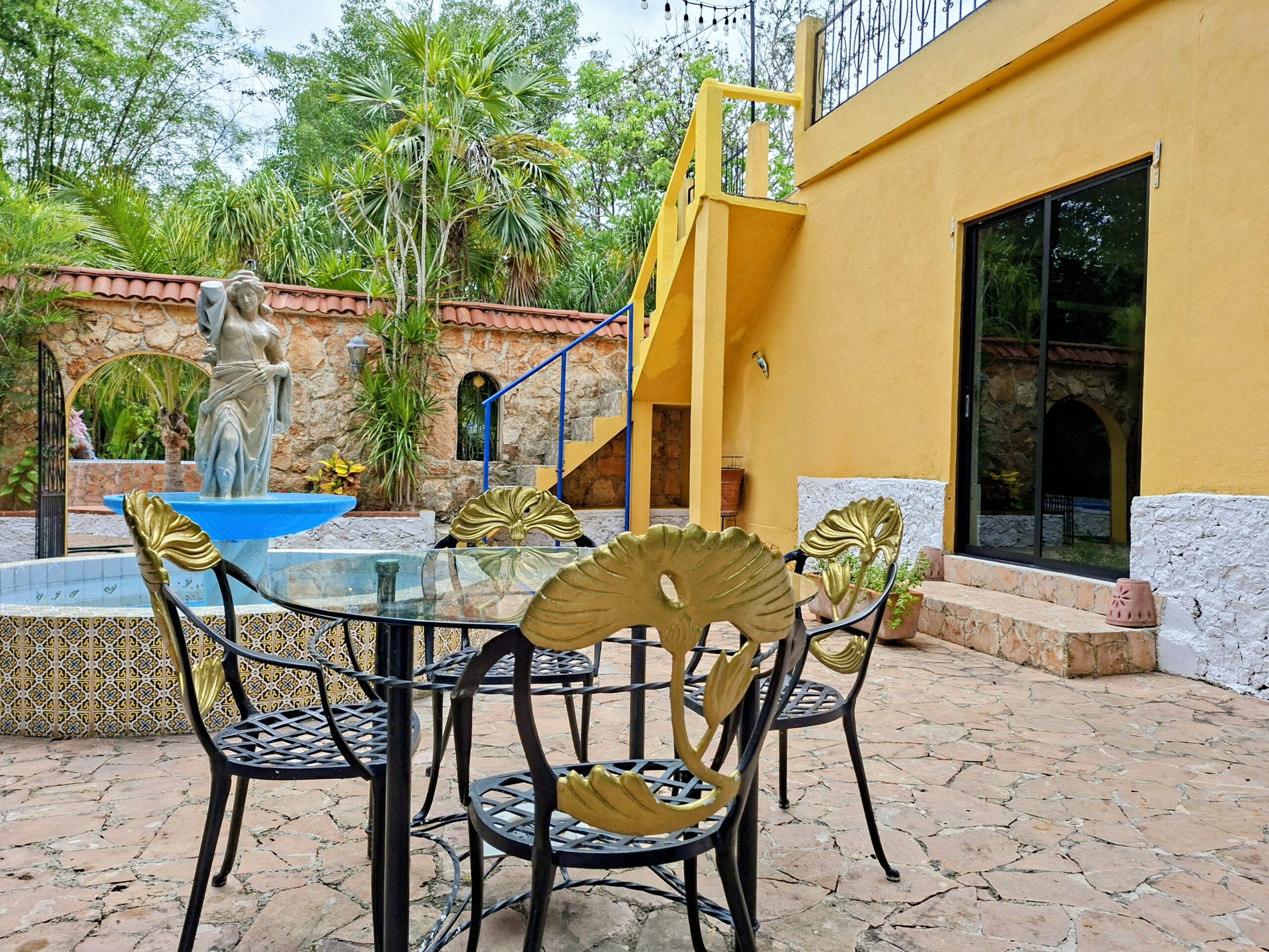 Outdoor courtyard with a fountain statue in a blue basin, surrounded by tropical plants and a stone wall behind a yellow building.