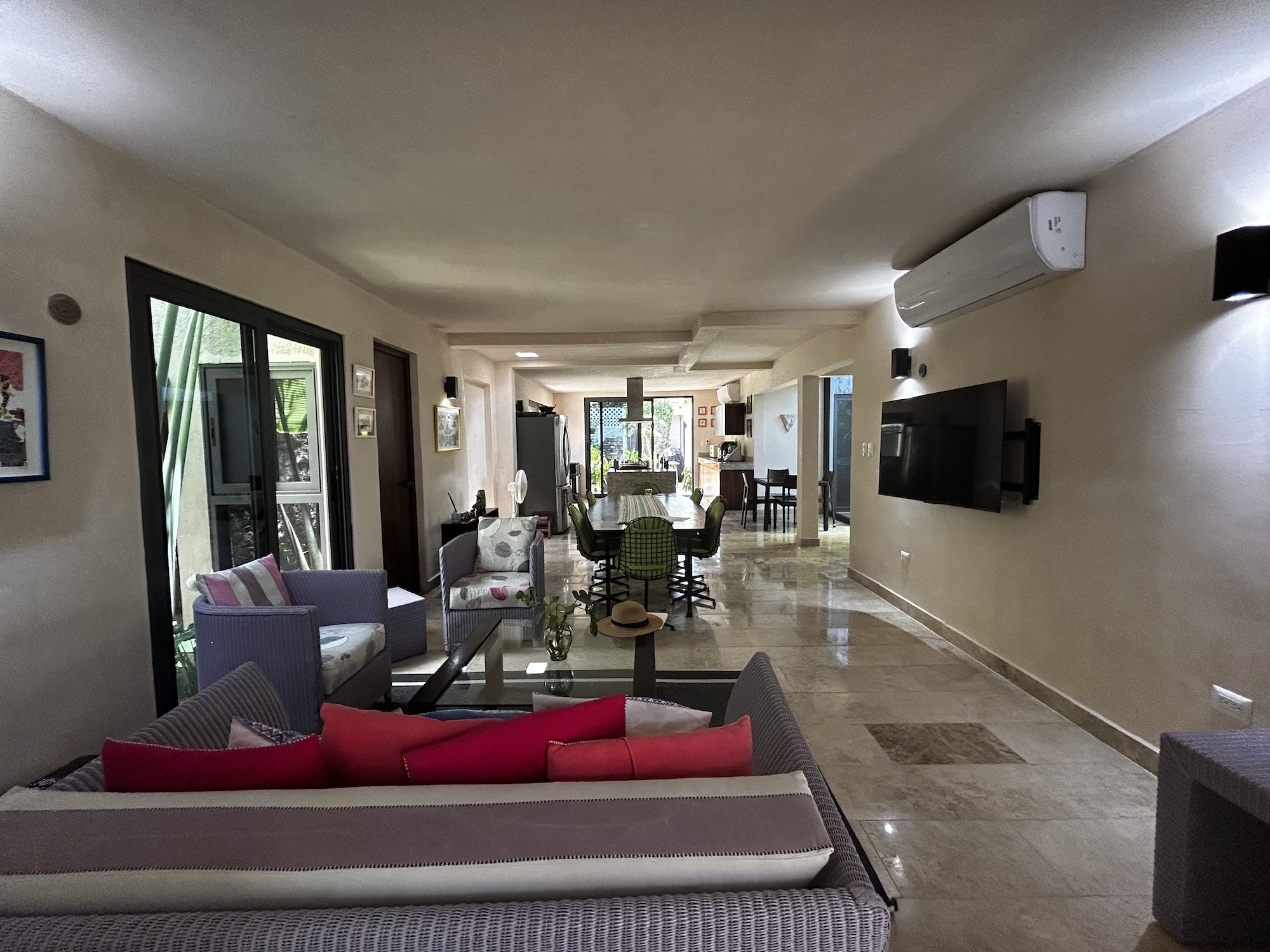 Spacious open-plan living area with wicker sofa and bright red cushions, a wall-mounted TV, sliding glass doors to the left, and a long dining table in the back.