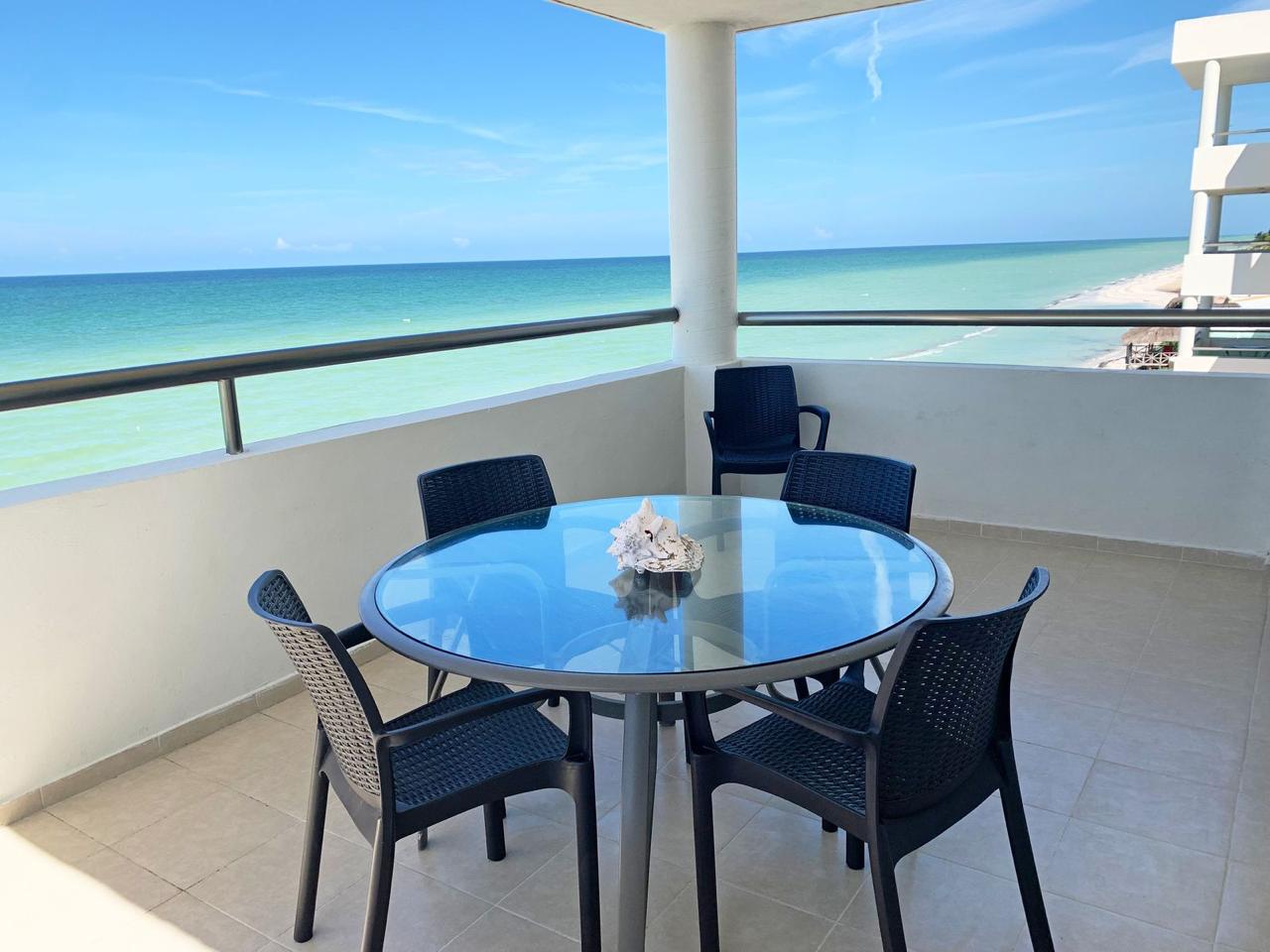 Coastal balcony with a round glass table and four black wicker chairs, overlooking turquoise sea and blue sky; shell centerpiece on the table.