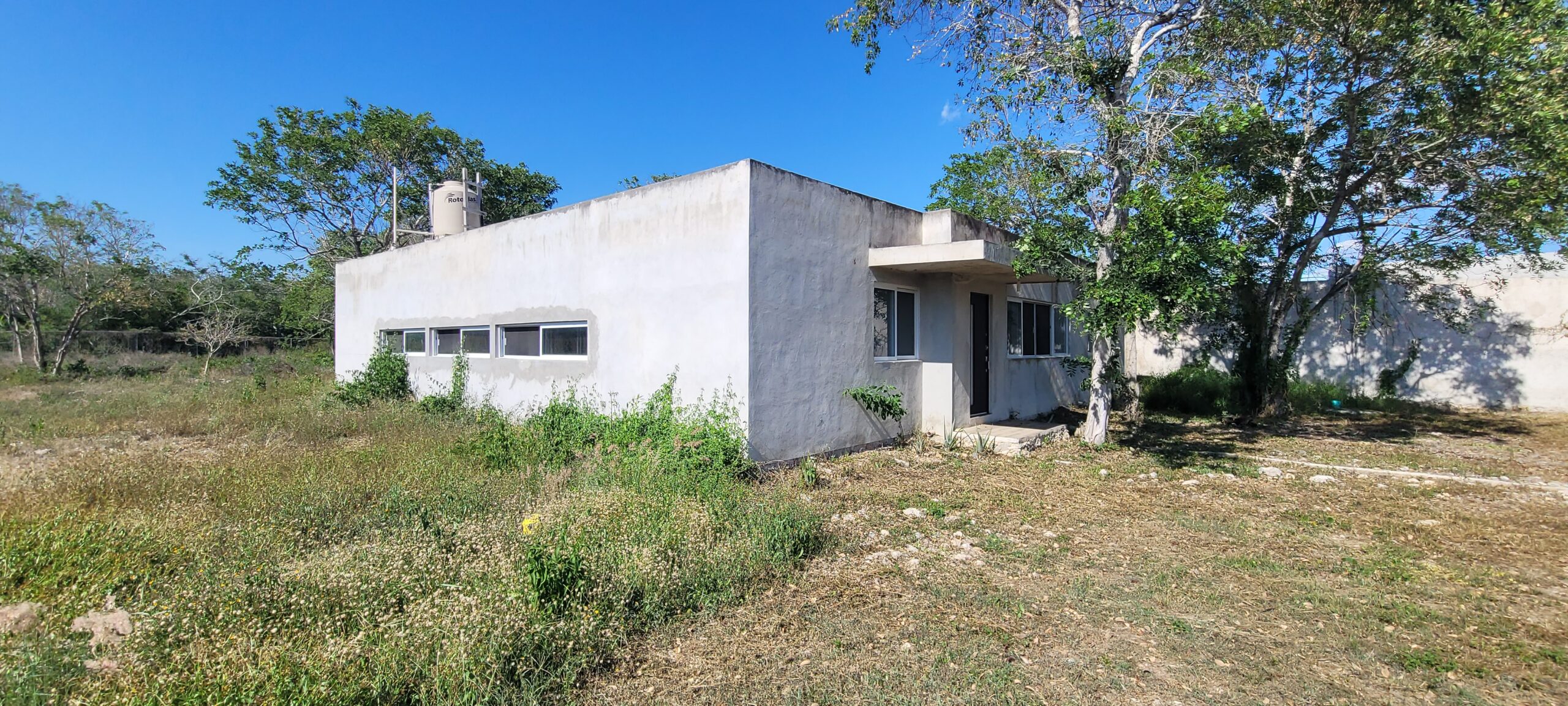 White single story concrete house with a flat roof and small windows set in a dry grassy lot under a bright blue sky with trees nearby