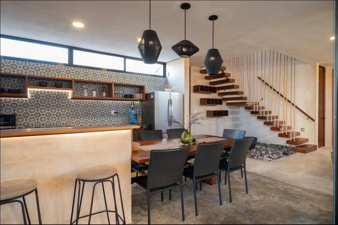 Open-concept kitchen and dining area with a patterned tile backsplash, a long wooden dining table and wicker chairs, and a floating wooden staircase with rope railings nearby.