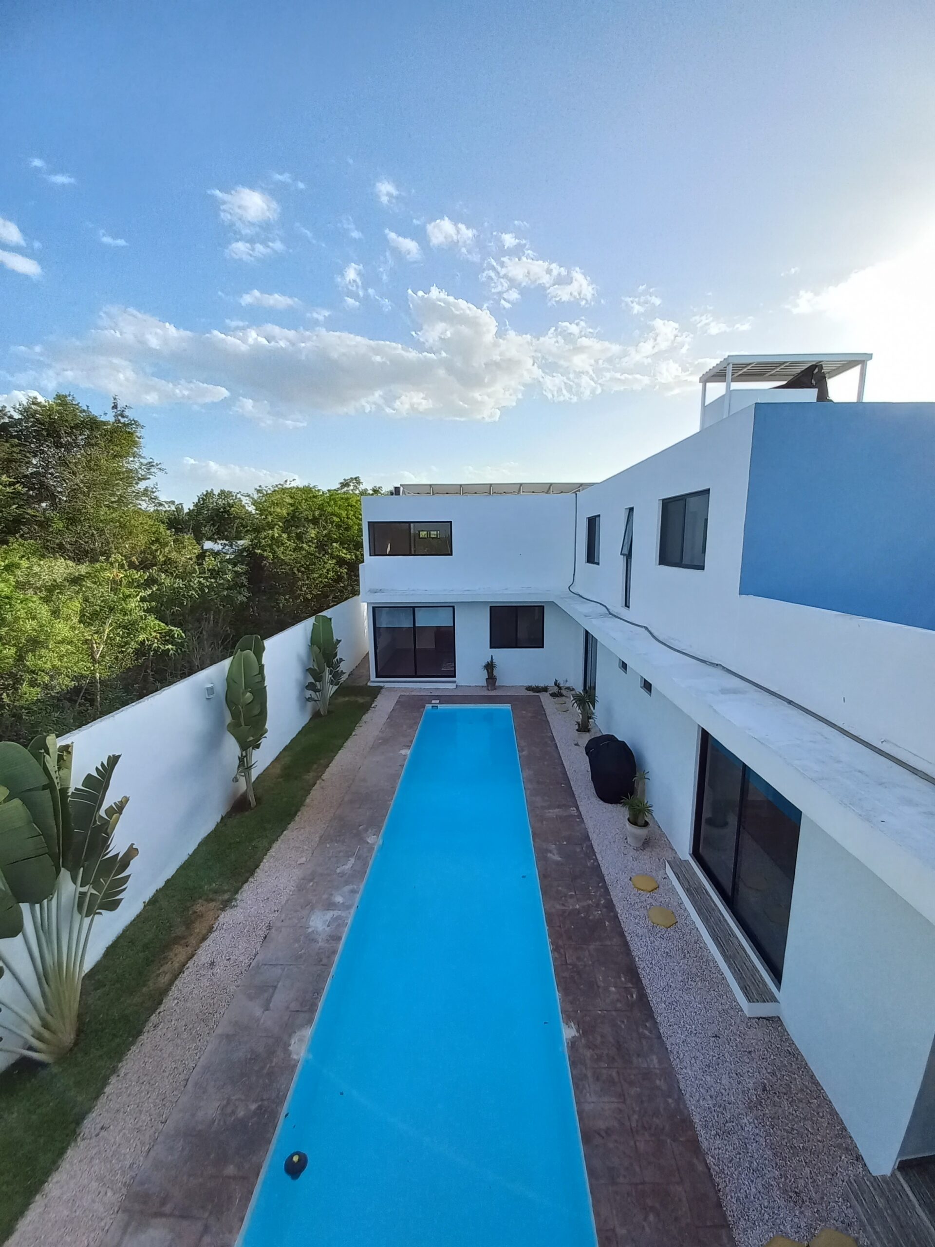 Aerial view of a long rectangular blue pool flanked by white modern buildings and green trees under a clear sky, courtyard with plants nearby.