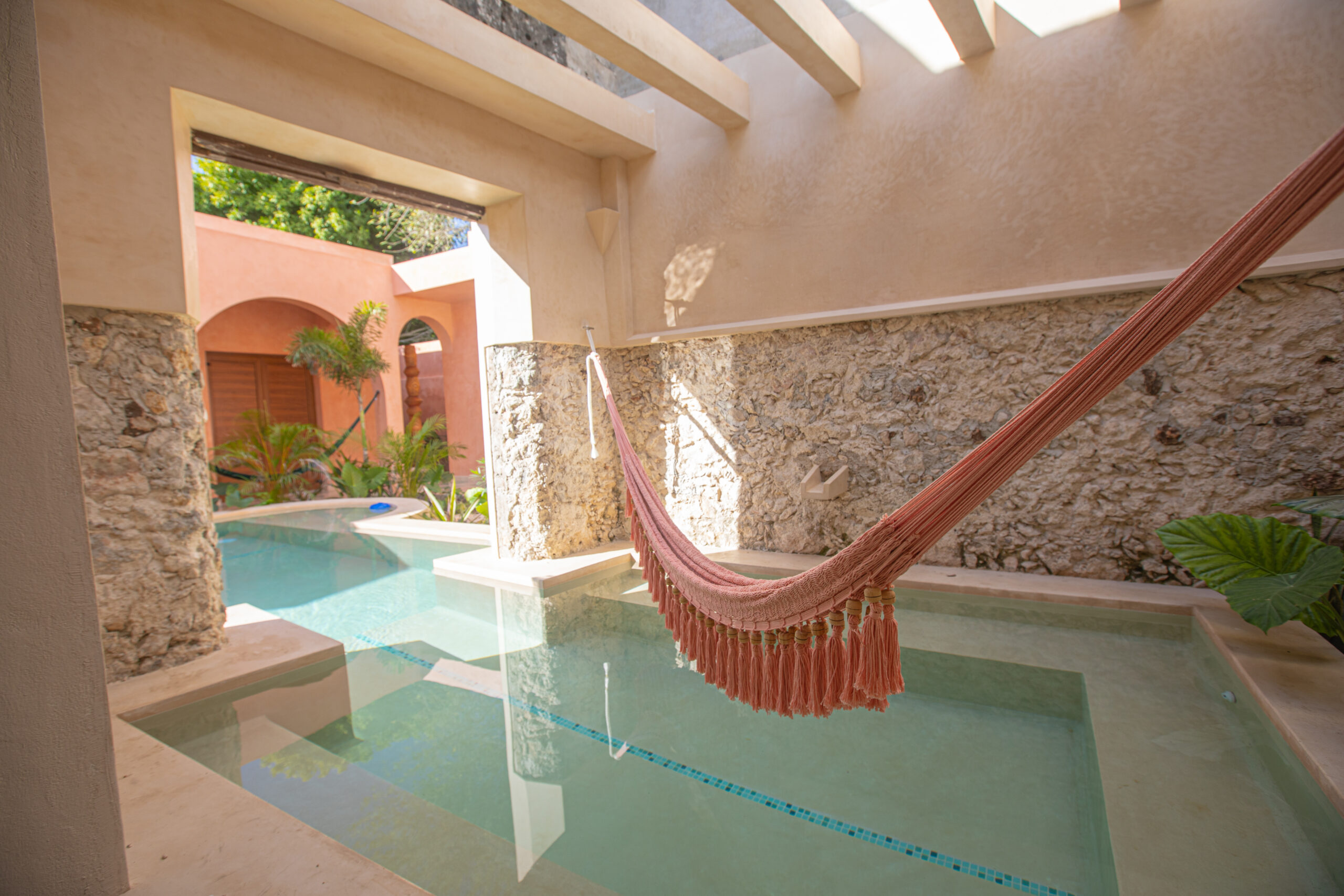 Sunlit poolside courtyard with a pink woven hammock strung between stone walls and arches