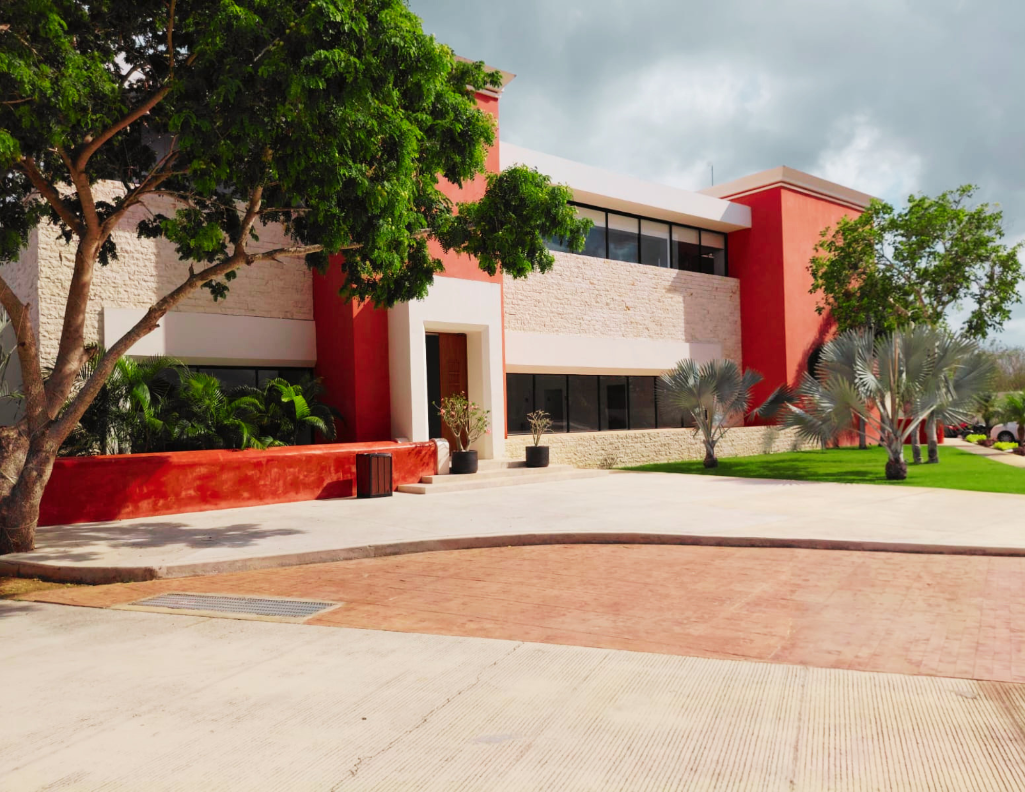 Modern beige and red building with large windows surrounded by trees and a paved courtyard
