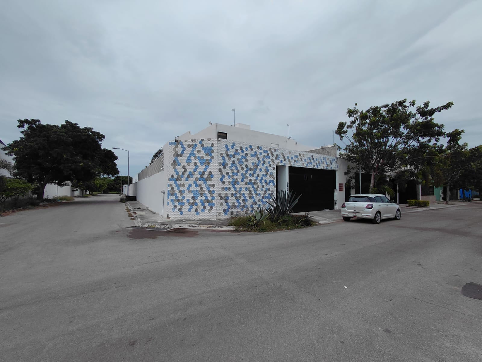White building with blue mosaic wall at a street corner open garage door and a parked white hatchback cloudy sky above