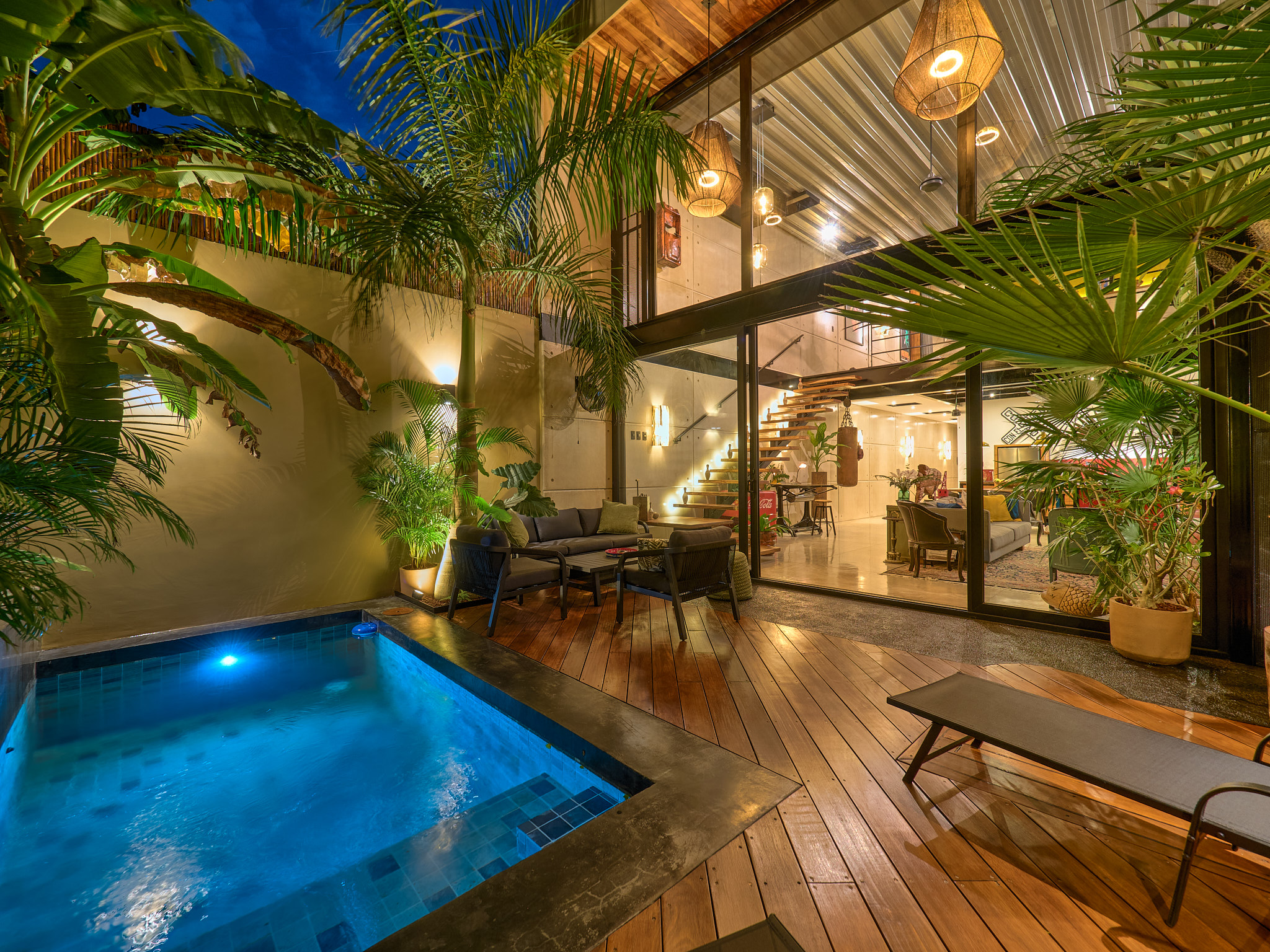 Indoor tropical courtyard at night pool wooden deck lush palms and glass walled living area with warm pendant lights