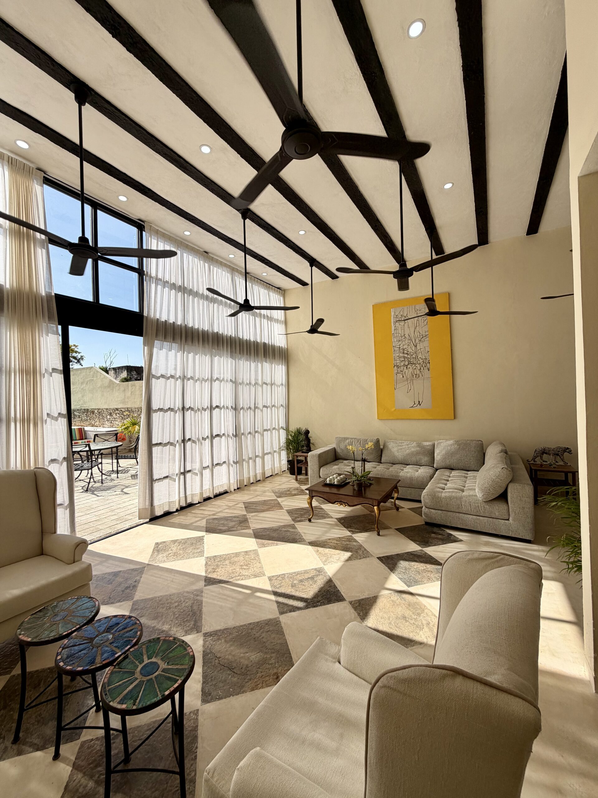 Bright living room with multiple ceiling fans large windows with sheer curtains and a yellow framed wall art piece above a beige sectional sofa on a checkered floor outside patio nearby