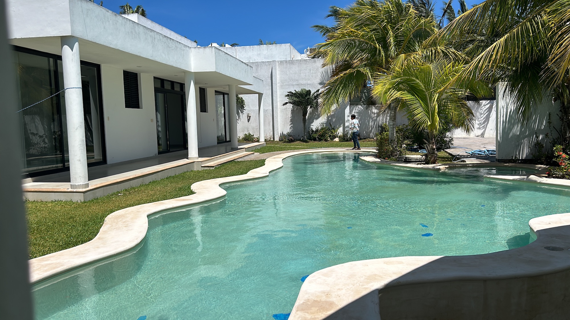 Modern white villa with a curved pool palm trees and a person standing by the water in a sunny courtyard