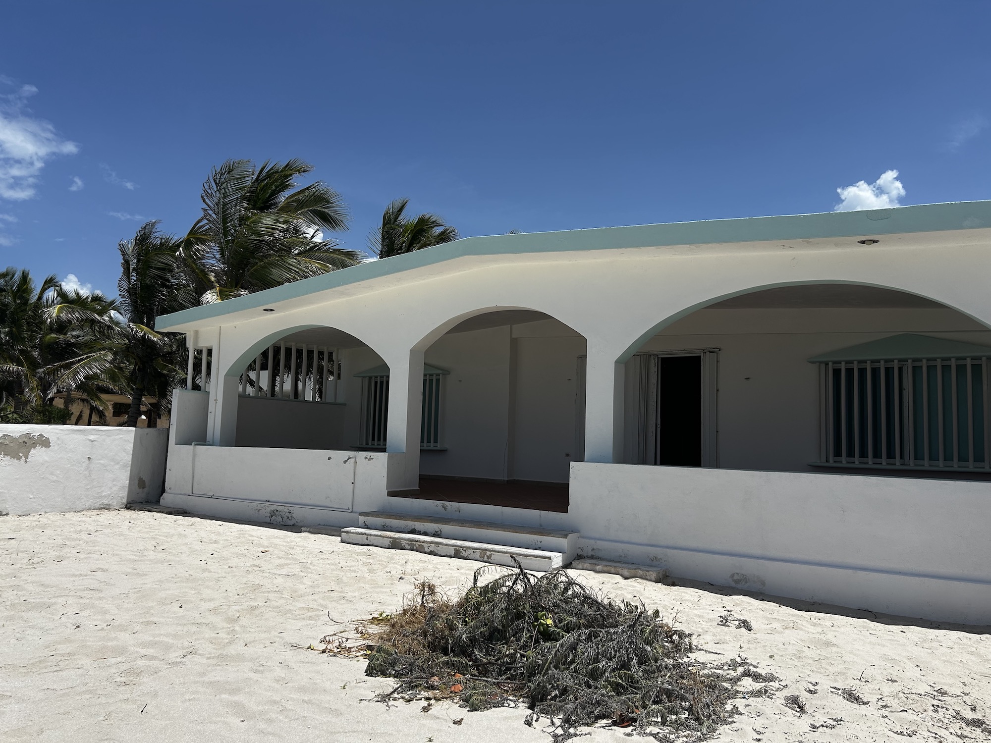 White building with arched colonnade on a sandy beach palm trees lining the left and a clear blue sky above