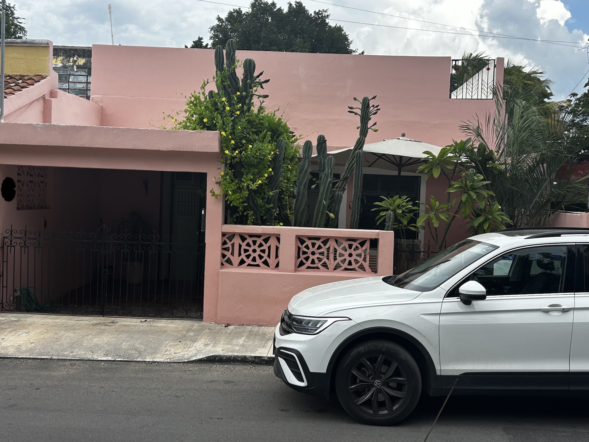 Pink stucco house with decorative lattice fence; tall cacti and greenery in front, and a white SUV parked along the curb.