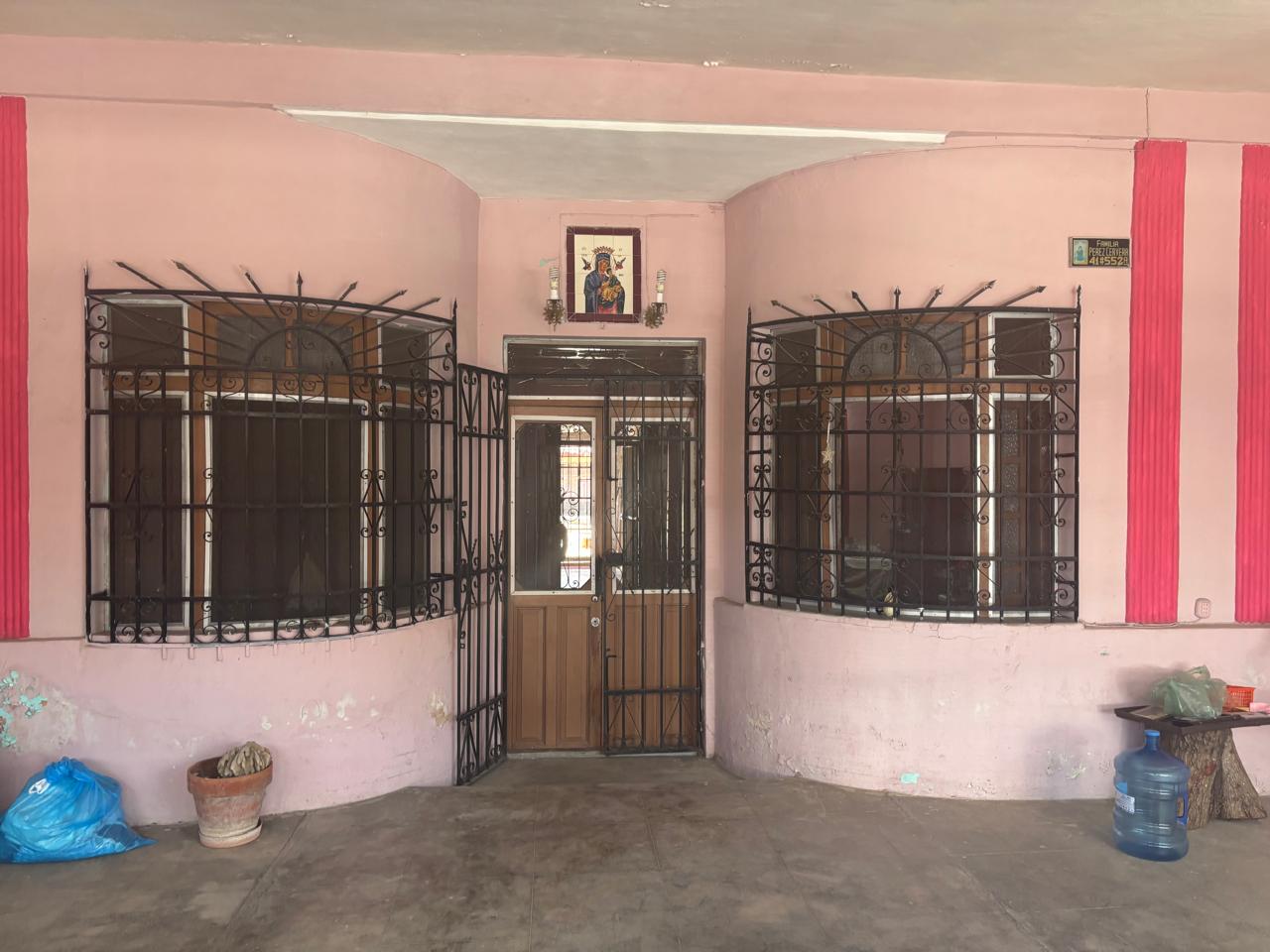 Pink façade with rounded window alcoves protected by decorative black wrought-iron bars and a central wooden door, small shrine above the door, items outside including a blue bag and a water jug nearby.