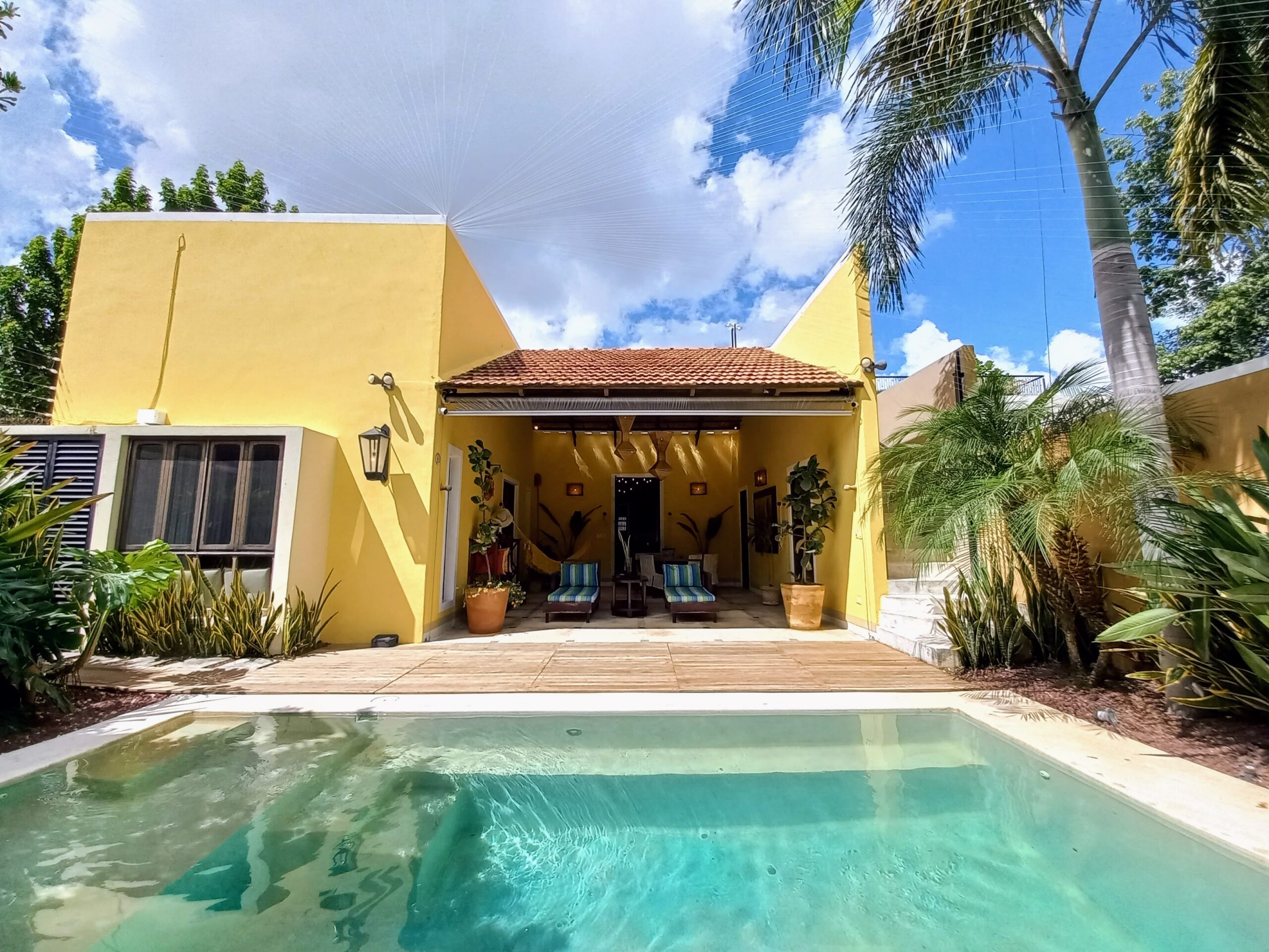 Yellow building with red-tiled roof over a shaded patio and lounge chairs, facing a swimming pool under a sunny sky with palm trees.