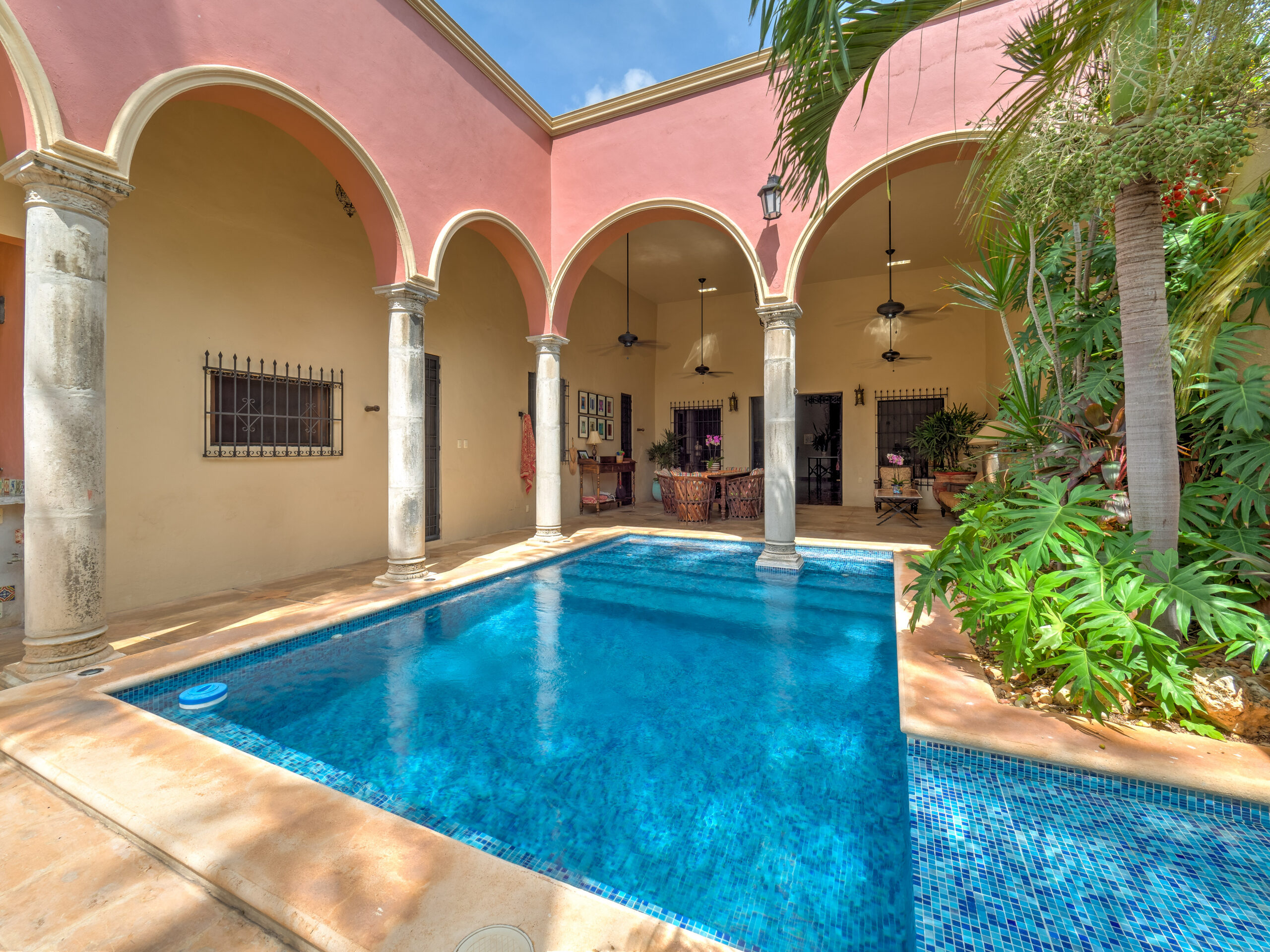 Open-air courtyard with pink arches, stone columns, and a blue-tiled pool, surrounded by tropical plants on the right.