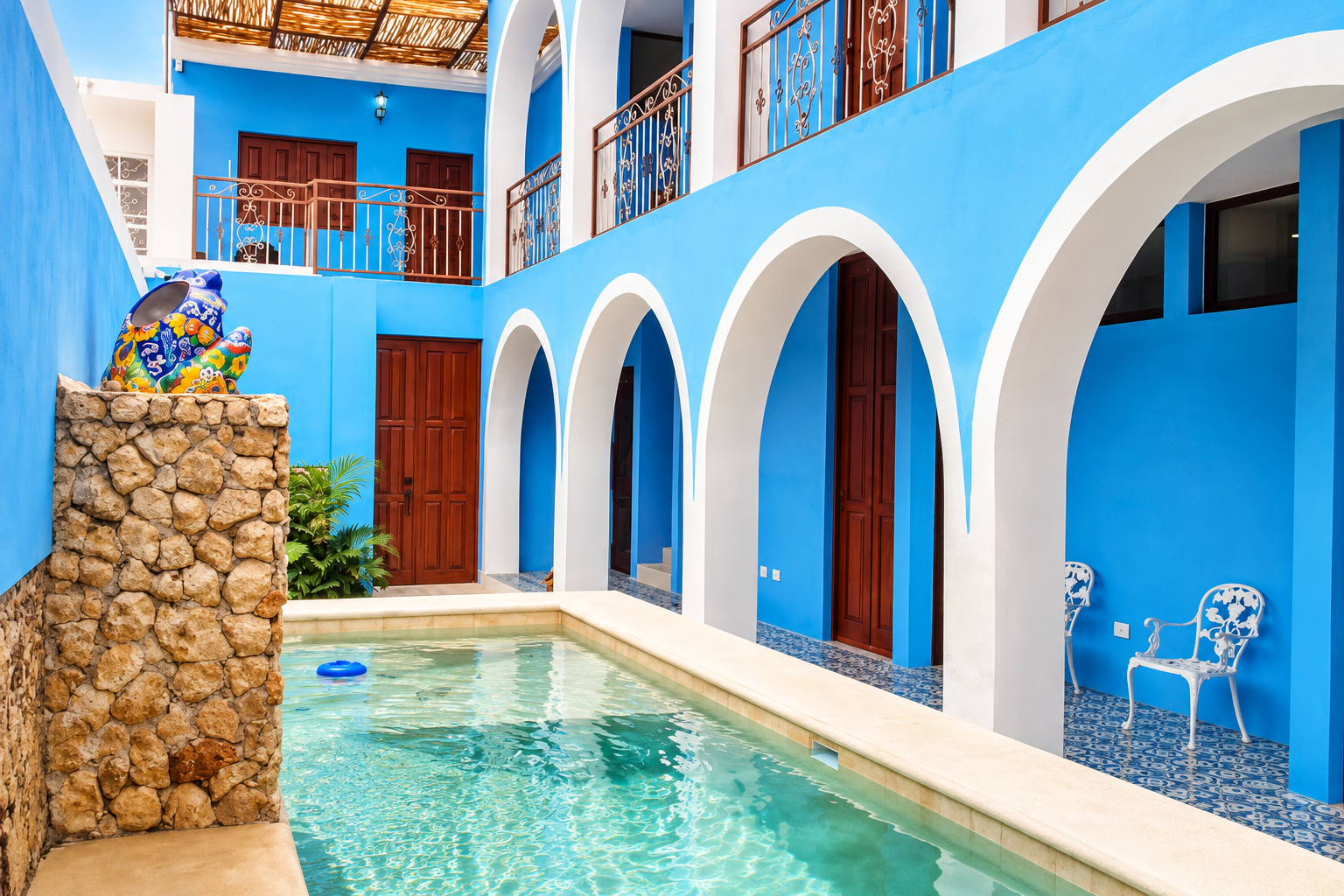 Blue-walled courtyard with white arches around a narrow pool, a colorful mosaic vase on a stone pedestal, and wooden doors.