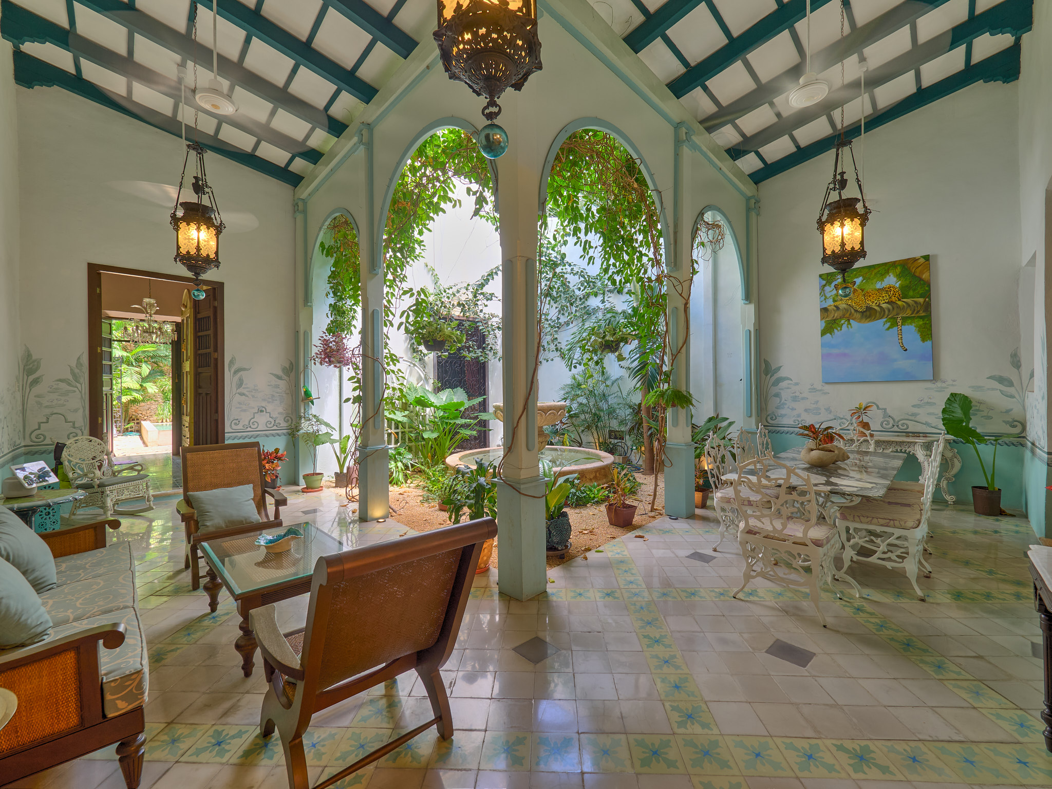 A sunlit covered veranda with teal arches, hanging lanterns, and lush potted plants surrounding white wrought-iron seating areas.