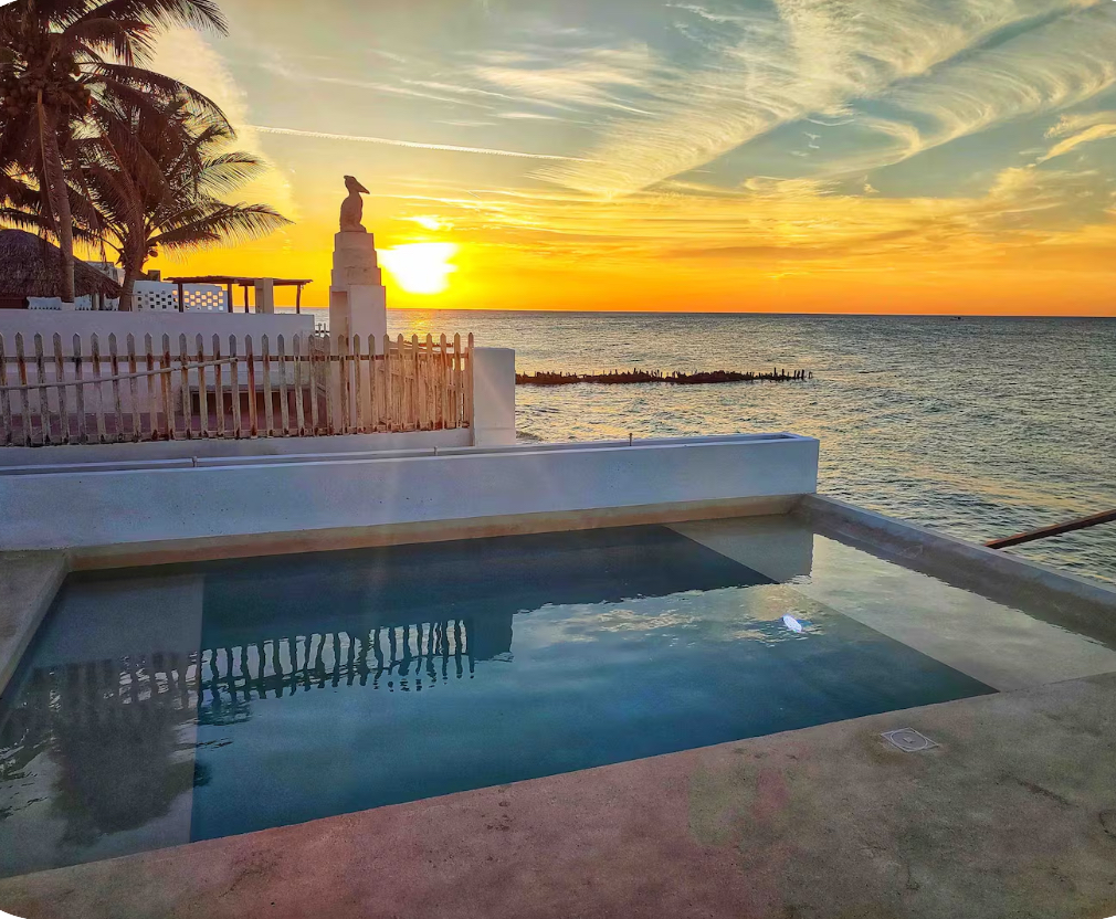 Sunset over the ocean visible beyond a white pool deck and low wall, with a fence and palm trees on the left.